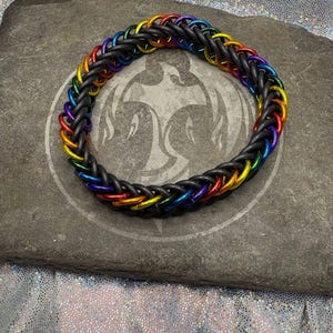 May include: A braided bracelet with a rainbow color scheme. The bracelet is made of black rubber and features colorful metal rings in red, orange, yellow, green, blue, and purple. The bracelet is displayed on a gray stone surface.