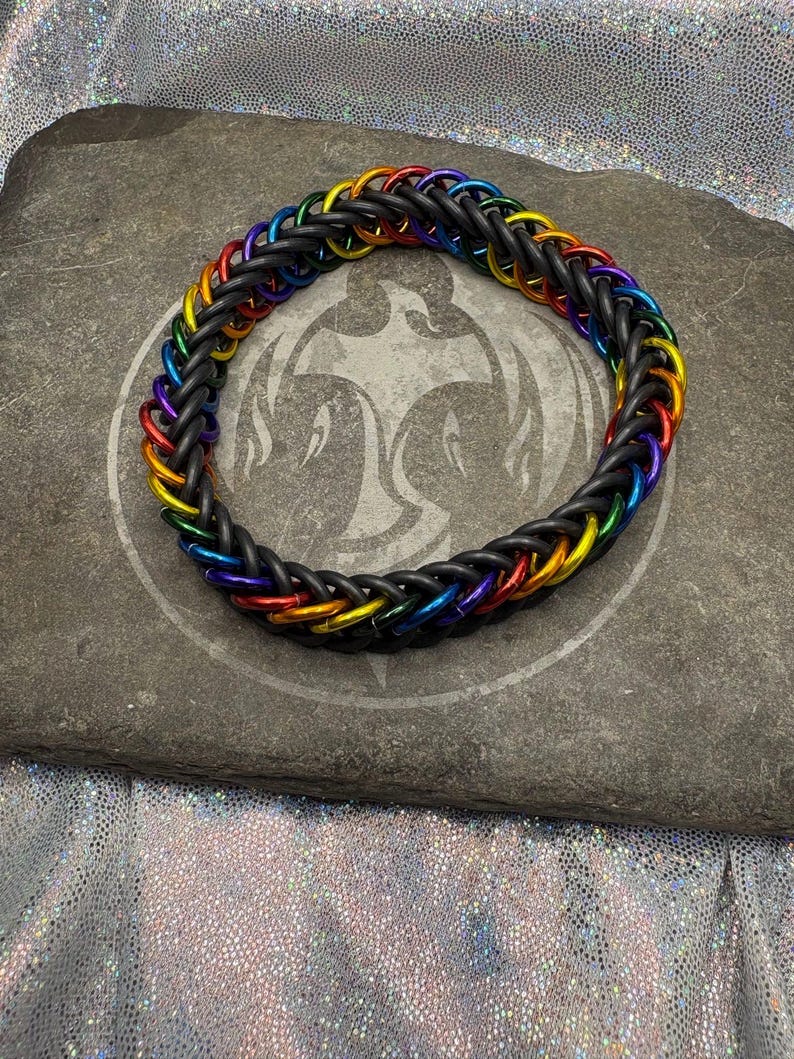 May include: A braided bracelet with a rainbow color scheme. The bracelet is made of black rubber and features colorful metal rings in red, orange, yellow, green, blue, and purple. The bracelet is displayed on a gray stone surface.