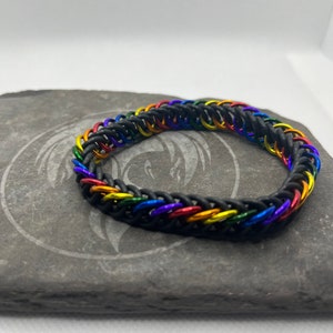 May include: A black chainmail bracelet with rainbow-colored links. The bracelet is made of small, interlocking metal rings in a pattern of red, orange, yellow, green, blue, and purple. The bracelet is displayed on a gray stone surface.