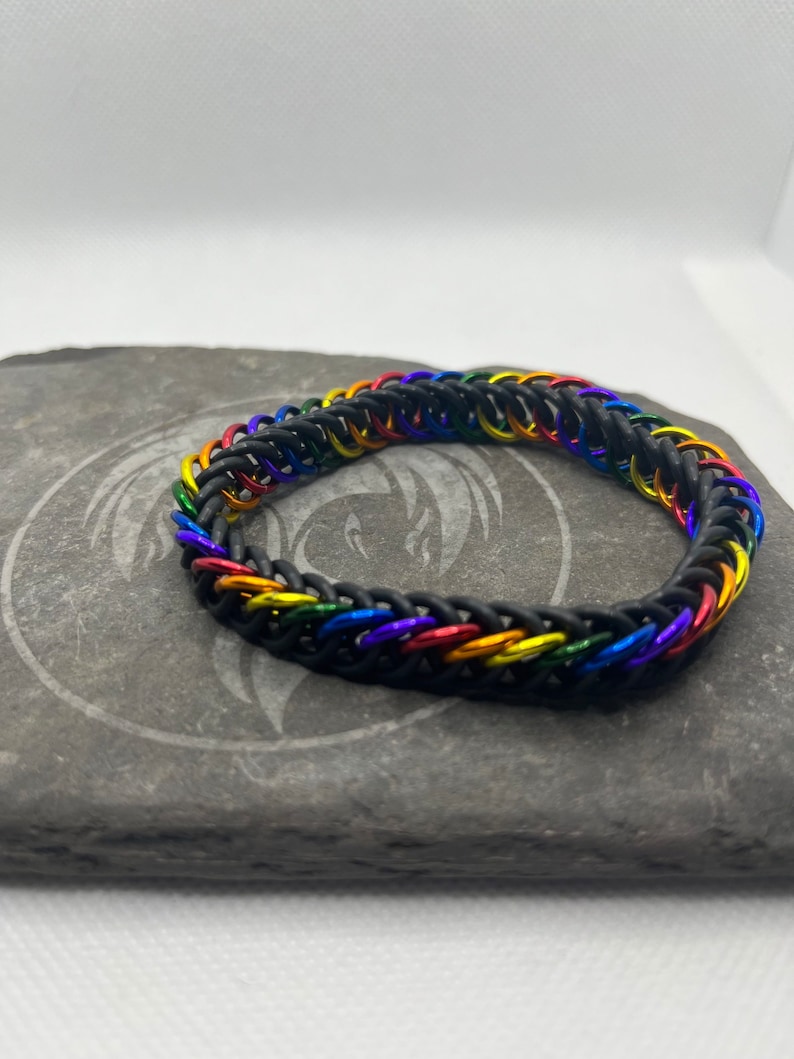 May include: A black chainmail bracelet with rainbow-colored links. The bracelet is made of small, interlocking metal rings in a pattern of red, orange, yellow, green, blue, and purple. The bracelet is displayed on a gray stone surface.