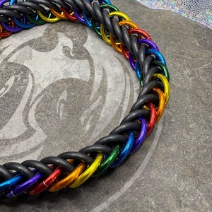 May include: A braided necklace featuring a rainbow of colors, including red, orange, yellow, green, blue, and purple, woven with black rubber tubing. The necklace is displayed on a gray surface.