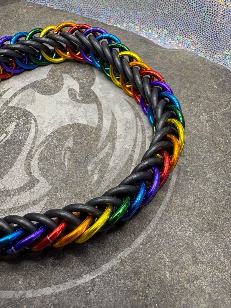 May include: A braided necklace featuring a rainbow of colors, including red, orange, yellow, green, blue, and purple, woven with black rubber tubing. The necklace is displayed on a gray surface.