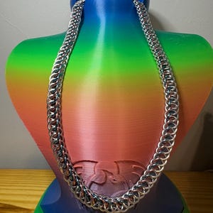 May include: A silver-toned chainmail necklace featuring a repeating interlocking ring pattern. The necklace is constructed from numerous small, circular metal links that create a flexible, textured, and continuous band that hangs in a U-shape.