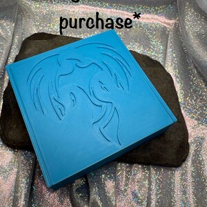 May include: A bright blue square gift box with a dragon design is displayed on a dark gray rock. The text "Free gift box with purchase*" and "*Color varies" are also visible.