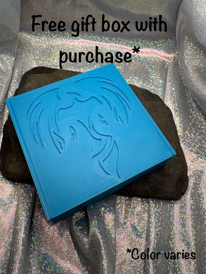 May include: A bright blue square gift box with a dragon design is displayed on a dark gray rock. The text "Free gift box with purchase*" and "*Color varies" are also visible.