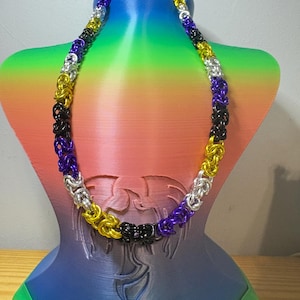 May include: A chainmail necklace constructed from interlocking metal rings in a repeating pattern of yellow, black, purple, and silver. The necklace features a Byzantine weave design, creating a textured, cylindrical appearance around the entire length.