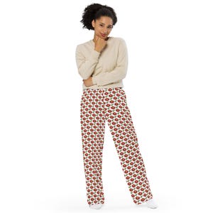 May include: A woman wearing a beige long-sleeve shirt and white pants with a red floral pattern. The pants have a wide leg design. The woman is standing in front of a white background.