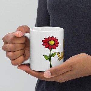 May include: White ceramic mug with a red daisy, yellow centre, and green stem and leaves. The mug has the words "I'm Sweet" in yellow script. The mug is held by a person.