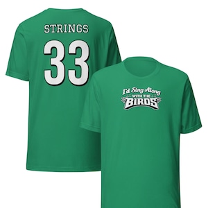 Puede incluir: Dos camisetas verdes. Una tiene "STRINGS" y el número "33" en blanco en la espalda. La otra tiene el texto "I'd Sing Along With The Birds" en un diseño blanco y negro.
