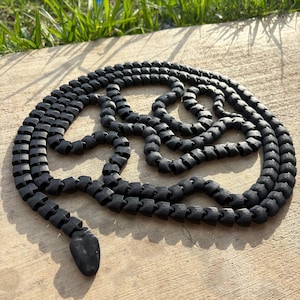 Puede incluir: Un juguete de serpiente negra articulada hecho de segmentos conectados. La serpiente está enrollada sobre una superficie de hormigón, con la cabeza en la parte inferior izquierda. El fondo incluye hierba verde y luz solar.