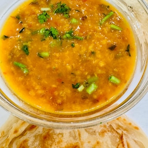 May include: A clear glass bowl filled with a vibrant orange-yellow soup, garnished with fresh green herbs and chopped green onions. The soup sits atop a stack of golden-brown, flaky flatbread.