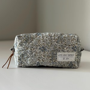 Peut inclure: Une trousse rectangulaire zippée avec un motif floral dense dans des tons de bleu, vert et marron. Une étiquette en tissu blanc est cousue sur le devant avec le texte noir WOOD AND THREADS BY JEN. Un cordon en cuir marron est attaché au zip.