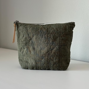 Puede incluir: Bolsa acolchada verde oliva con forma rectangular. La bolsa tiene una cremallera blanca y un tirador de cuero marrón. La tela tiene un patrón de cuadrícula creado por las costuras.