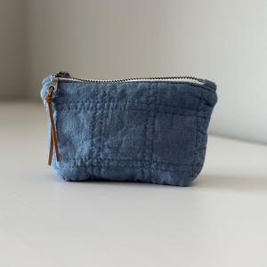 May include: A small, blue quilted pouch with a zip closure and a brown leather pull. The pouch is made of a soft, textured fabric and has a rectangular shape. The zip is silver-toned.