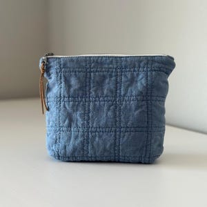 May include: A square, blue quilted pouch with a zipper closure and a brown leather pull. The pouch has a grid pattern and a white zipper. The pouch is sitting on a white surface.