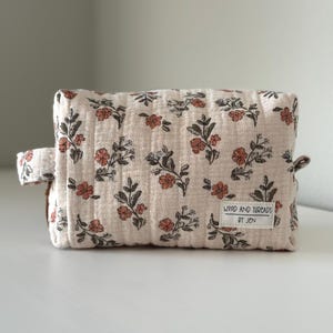 May include: A rectangular, beige floral patterned pouch with a handle on the side. The pouch features a repeating pattern of orange flowers and green leaves. The text "WOOD AND THREADS BY JEN" is printed on a small white tag.