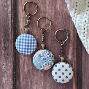 May include: Three circular keychains with bronze-toned metal rings and chains. One features a blue and white gingham pattern, one displays a blue floral print with red accents, and the third has a white background with blue polka dots.