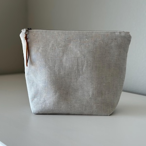 May include: A light gray linen pouch with a zipper closure and a brown leather pull. The pouch is rectangular with a slightly tapered shape. The fabric has a textured appearance.