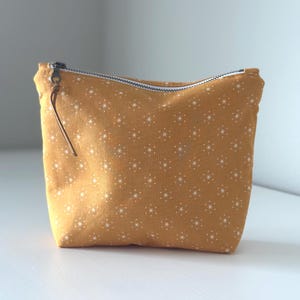 May include: Mustard-colored zippered pouch with a white starburst pattern. The pouch has a brown leather zipper pull and a silver zipper. The fabric appears to be cotton. The pouch is likely used for cosmetics or small items.