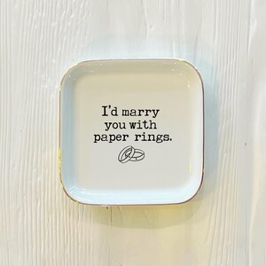 May include: White ceramic ring dish with a gold rim. The dish has the text "I'd marry you with paper rings." printed on it, with a simple illustration of two paper rings.