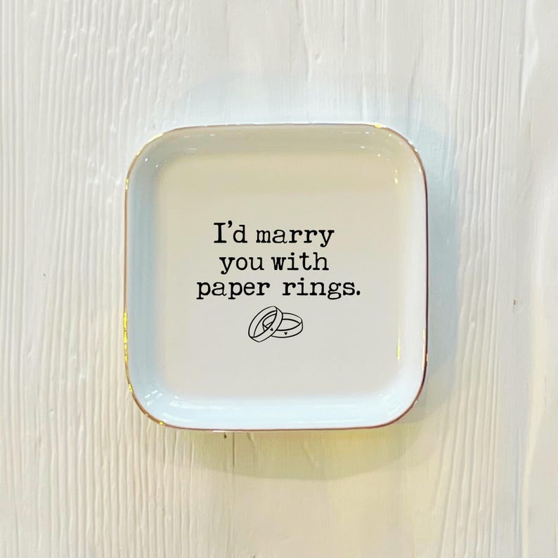 May include: White ceramic ring dish with a gold rim. The dish has the text "I'd marry you with paper rings." printed on it, with a simple illustration of two paper rings.