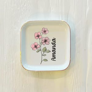 May include: White ceramic trinket tray with a gold rim. The tray features a pink floral design and the name "Amanda" written in black.