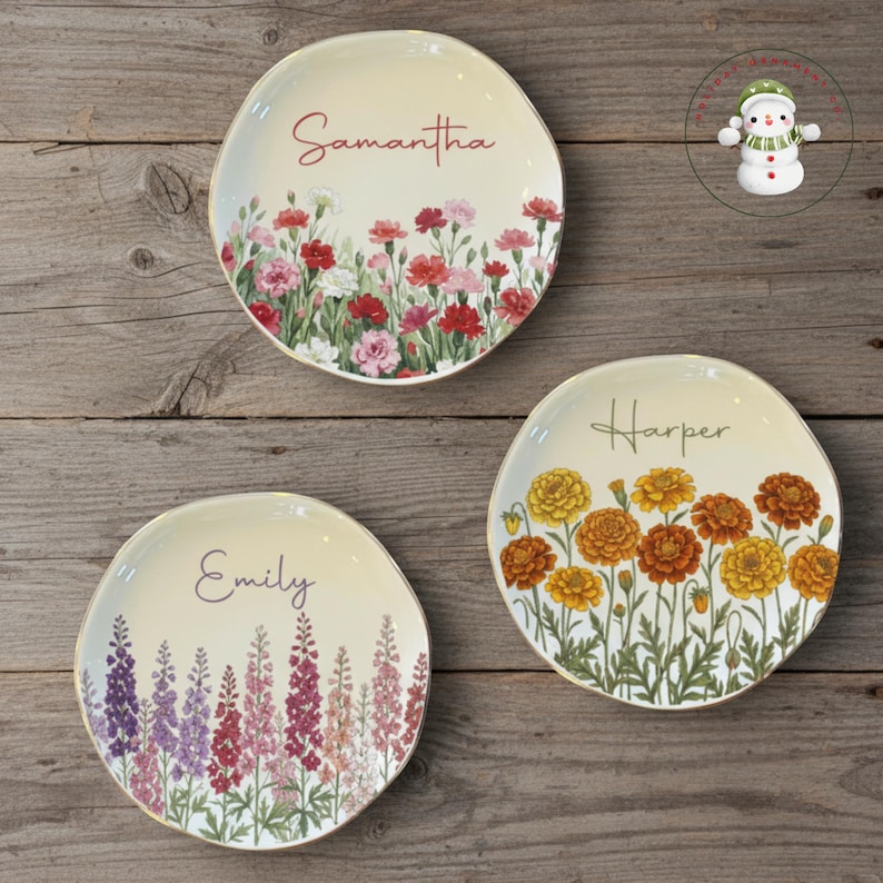 May include: Three decorative ceramic plates with floral designs and gold rims. Each plate features a different flower arrangement and a name: "Samantha", "Emily", and "Harper". The plates are on a wooden surface.