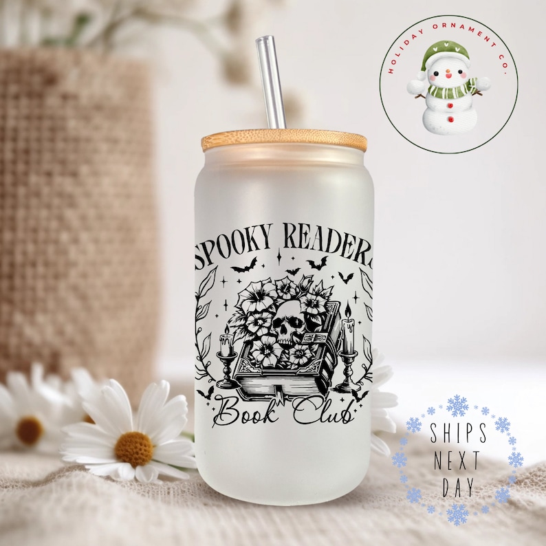 Puede incluir: Un vaso de vidrio esmerilado con un dise&ntilde;o en blanco y negro de una calavera, flores y un libro con el texto "Spooky Reader Book Club". El vaso tiene una tapa de bamb&uacute; y una pajita transparente.