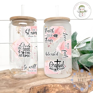 May include: Two glass beverage cups with wooden lids and straws. One is clear, the other frosted. Both feature inspirational quotes in black and pink, including "God is Good" and "Faith Over Fear". A snowman logo is visible.
