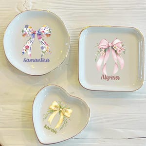 Puede incluir: Tres platos de cerámica para joyas con ribete dorado. Los platos tienen forma de corazón, cuadrado y círculo. Cada plato presenta un lazo de acuarela con detalles florales y un nombre personalizado: Samantha, Alyssa y Sarah.