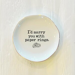 May include: White ceramic ring dish with a gold rim. The dish has the text "I'd marry you with paper rings." and a simple illustration of two rings.
