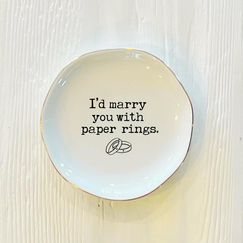 May include: White ceramic ring dish with a gold rim. The dish has the text "I'd marry you with paper rings." and a simple illustration of two rings.