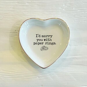 May include: White heart-shaped ceramic ring dish with a gold rim. The dish has the text "I'd marry you with paper rings." printed on it, with a small illustration of two paper rings.