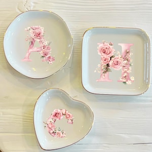 Puede incluir: Tres platos decorativos de cerámica de diferentes formas: redondo, cuadrado y en forma de corazón. Cada plato presenta una letra inicial rosa, adornada con rosas rosas y detalles florales. Los bordes de los platos son dorados.