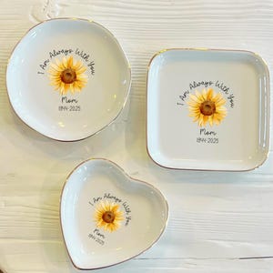 May include: Three ceramic ring dishes with gold trim. Each dish features a sunflower with the text "I Am Always With You Mom 1944-2025". The dishes are shaped like a circle, a square, and a heart.