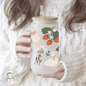 May include: A frosted glass water bottle with a bamboo lid and a straw. The bottle has a whimsical design featuring white ducks, red strawberries, and green leaves.