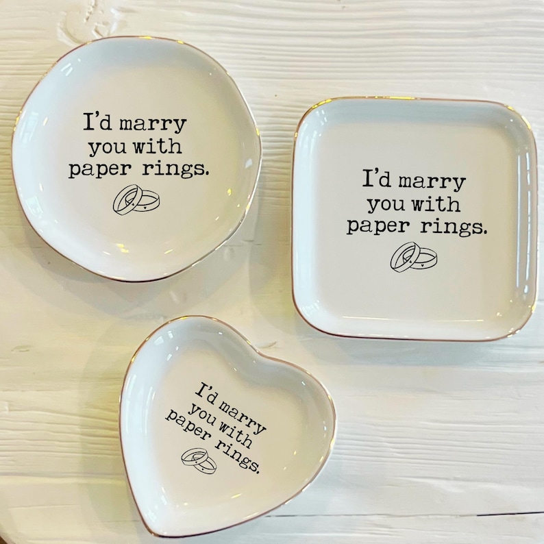 May include: Three white ceramic ring dishes with gold trim. Each dish has the text "I'd marry you with paper rings." and a small illustration of two rings. The dishes are in the shapes of a circle, a square, and a heart.