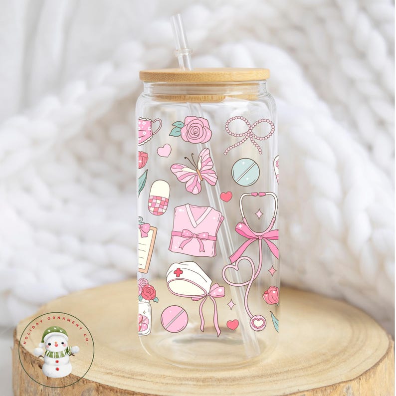 May include: A clear glass tumbler with a bamboo lid and straw, decorated with pink medical-themed illustrations. The design includes a stethoscope, scrubs, pills, and a butterfly. The tumbler sits on a wooden surface.