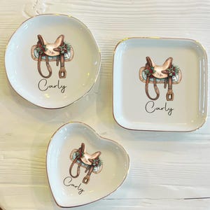 Op de afbeelding: Drie witte keramische sieradenschaaltjes in ronde, vierkante en hartvormige uitvoeringen. Elk schaaltje heeft een aquarel illustratie van een paardenzadel met feestelijk groen en de naam "Carly" in script. De randen van de schaaltjes zijn goudkleurig.