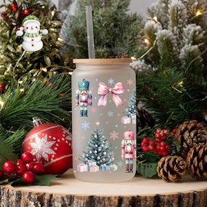 May include: Frosted glass tumbler with a wooden lid and straw. The tumbler is decorated with a Christmas theme, featuring nutcrackers, a pink bow, Christmas trees, snowflakes, and gift boxes. The tumbler is surrounded by Christmas decorations.
