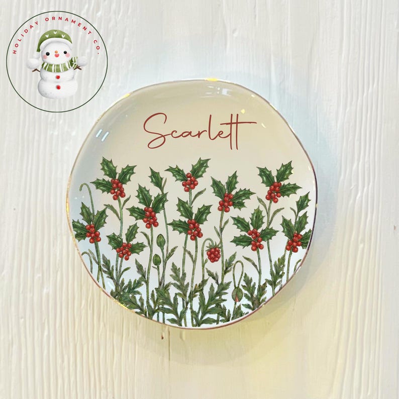 May include: A white ceramic plate with a gold rim, featuring a holly and berry design. The name "Scarlett" is written in red script across the top. A snowman logo is in the upper left corner.