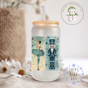 May include: A frosted glass tumbler with a wooden lid and a straw. The tumbler features a vintage-style illustration of a ballerina and a nutcracker on a blue background with snowflakes. The text "Holiday Ornament Co." is visible in a circle at the top of the image.