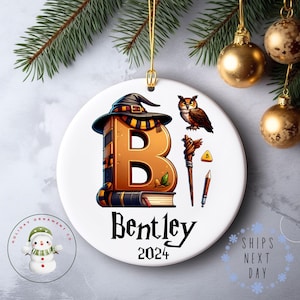 May include: A round white ceramic ornament featuring a large orange letter B designed as a book with a wizard hat on top. Beside it are an owl, a wooden wand, a small yellow triangle, and a pencil. The name Bentley and the year 2024 are printed below.