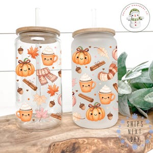 Peut inclure: Deux gobelets en verre avec couvercles et pailles en bambou. L'un est transparent, l'autre givré. Les deux sont décorés d'illustrations sur le thème de l'automne, notamment des citrouilles, des tasses à café, des bâtons de cannelle et des feuilles. Les gobelets sont posés sur une surface en bois.