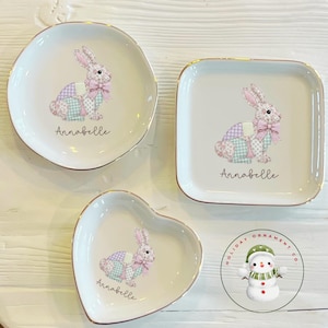 以下が含まれることがあります： 金色の縁取りが施された3つの白いセラミック皿。各皿には、ピンクのリボンと「アナベル」という名前が付いたパステルパッチワークのウサギのデザインが施されています。お皿は丸、四角、ハート型です。