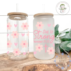 May include: Two frosted glass tumblers with bamboo lids and straws. One tumbler has a pink floral and heart design. The other tumbler has a pink floral design with the text "She is Strong Proverbs 31:25" and a cross.