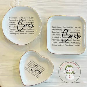 May include: Three white ceramic trinket dishes with gold-toned rims, including one round, one square, and one heart-shaped dish. Each dish features black printed text listing qualities like Organizer, Instructor, Guide, and the word Coach in script.