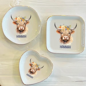 May include: Three decorative ceramic dishes in different shapes: round, square, and heart-shaped. Each dish features a watercolor illustration of a Highland cow with a floral crown and the name "HANNAH" in navy blue. The dishes have gold rims.