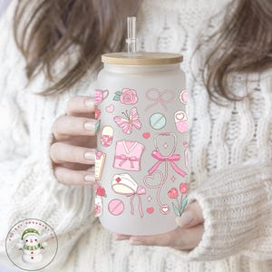 May include: Frosted glass tumbler with a bamboo lid and straw. The tumbler is decorated with pink medical-themed illustrations, including a stethoscope, pills, and a nurse's cap. The design is repeated around the entire glass.
