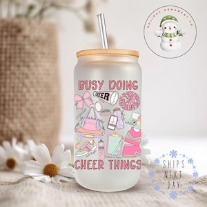 May include: A frosted glass tumbler with a bamboo lid and a straw. The tumbler has a pink and white design with the text "Busy Doing Cheer Things". The design includes cheerleading pom-poms, a backpack, makeup, and other cheerleading-related items.